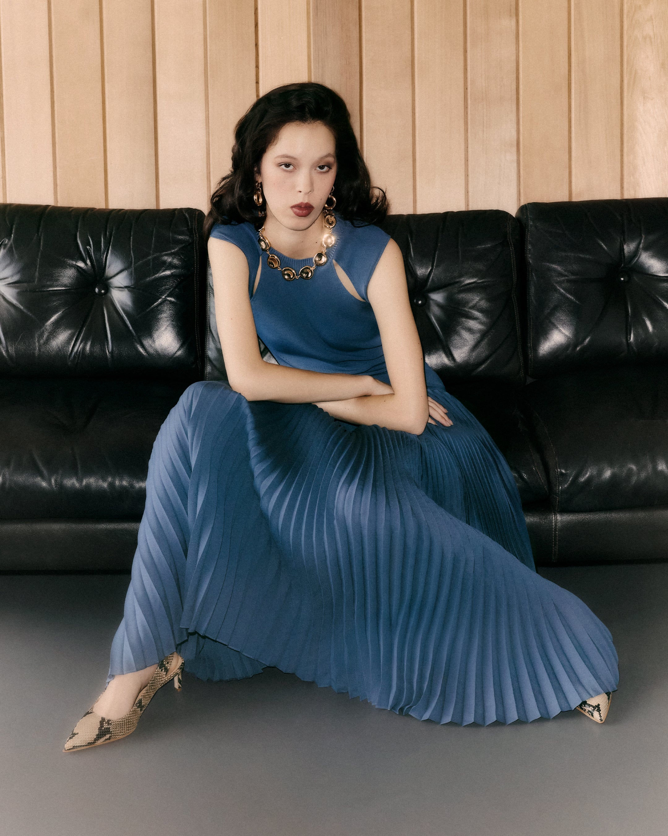 A woman wearing a sleeveless blue pleated maxi dress with cutout details and gold jewelry, sitting on a black leather sofa against a wood-paneled wall.