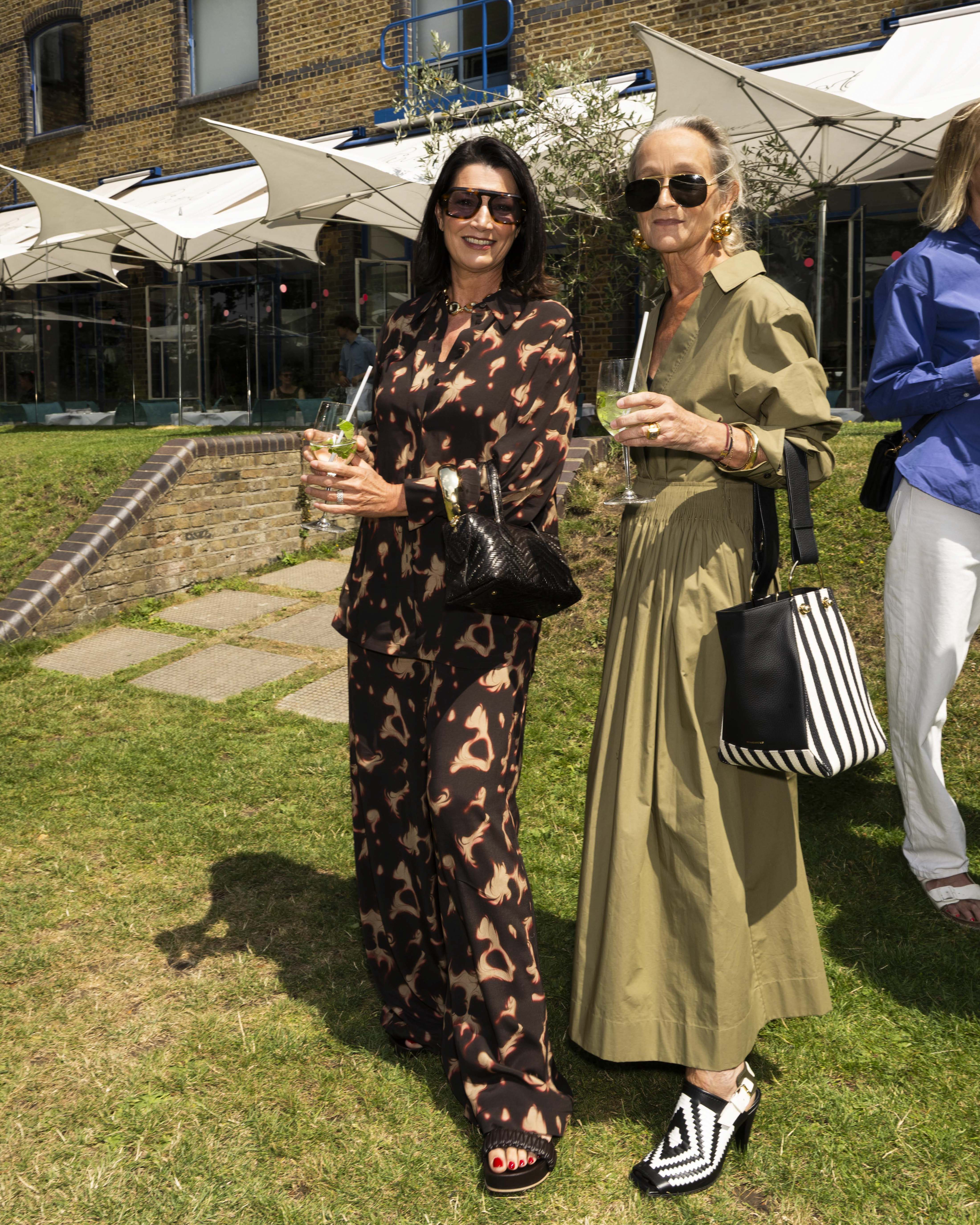 Barbara Campos, Lucinda Chambers posing outdoors on a grassy lawn at a summer event. The woman on the left wears a brown and black print co-ord and holds a black handbag. The woman on right in a khaki green dress.