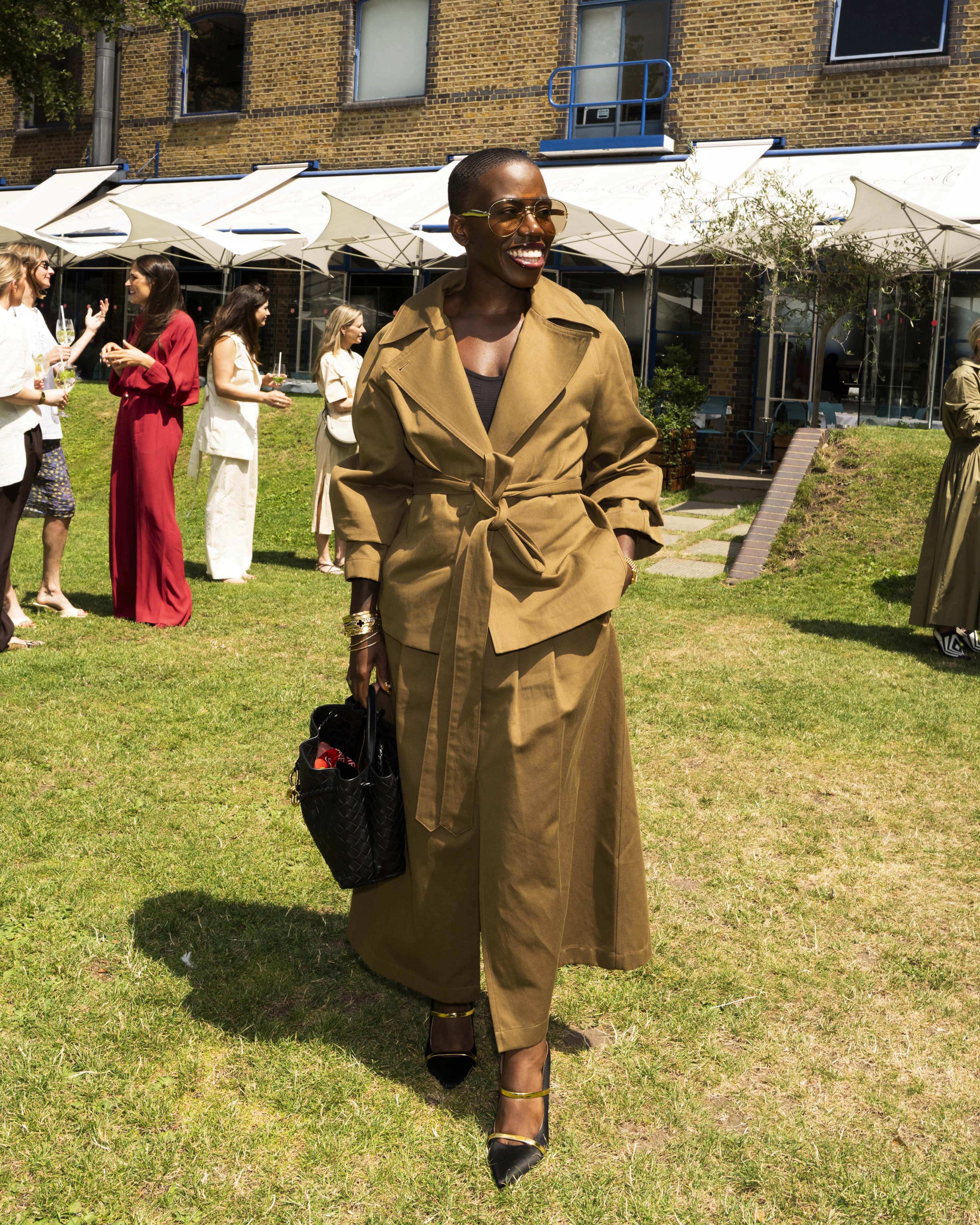 Candice Brathwaite at a summer event wearing a camel brown belted trench coat and matching trousers, holding a black handbag. In the background, people gather near white umbrellas in front of a brick building.