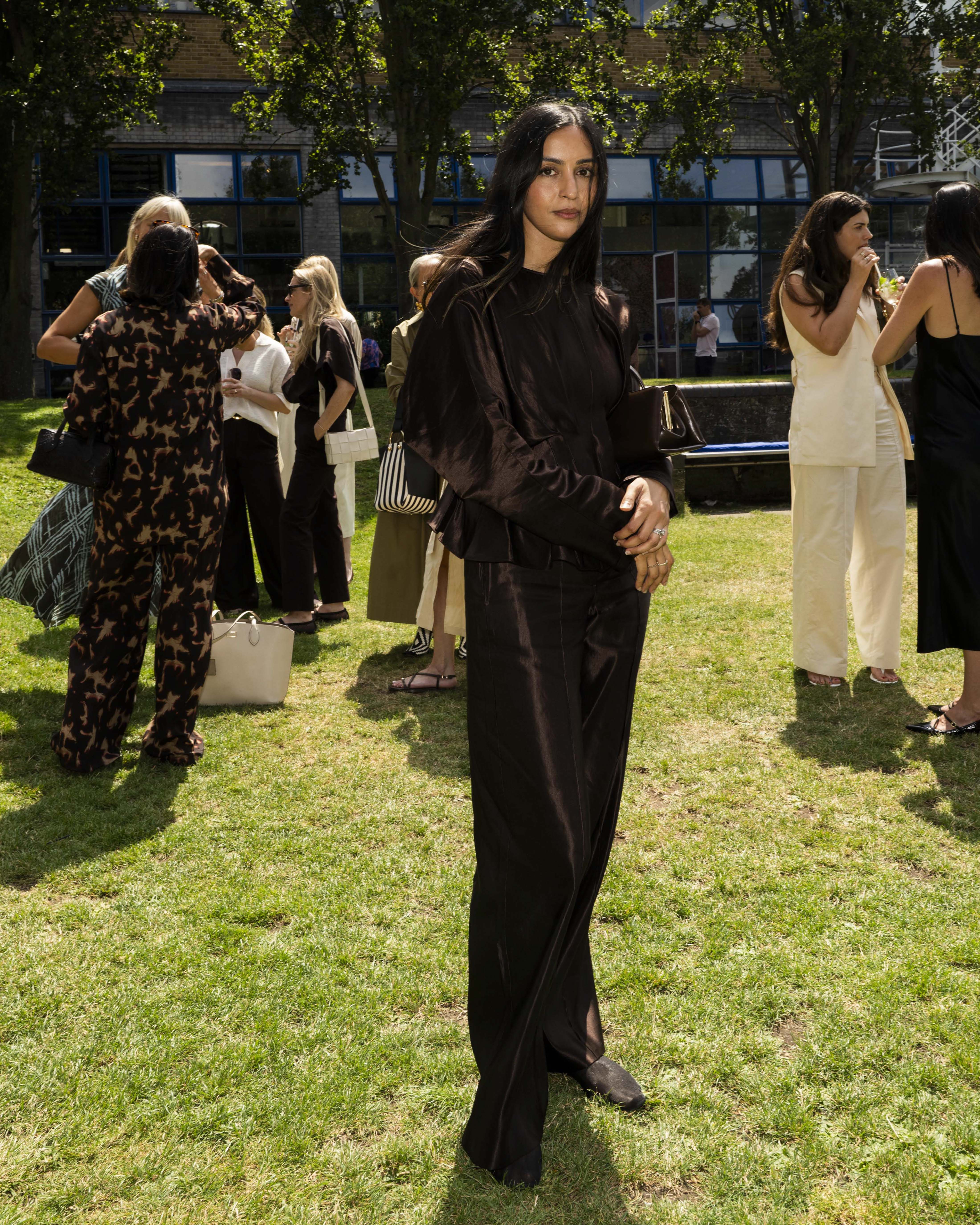 Ume Romaan stands at a summer event wearing a brown satin long-sleeve top and matching wide-leg trousers. Other people in casual and patterned outfits are visible in the background among trees and buildings.