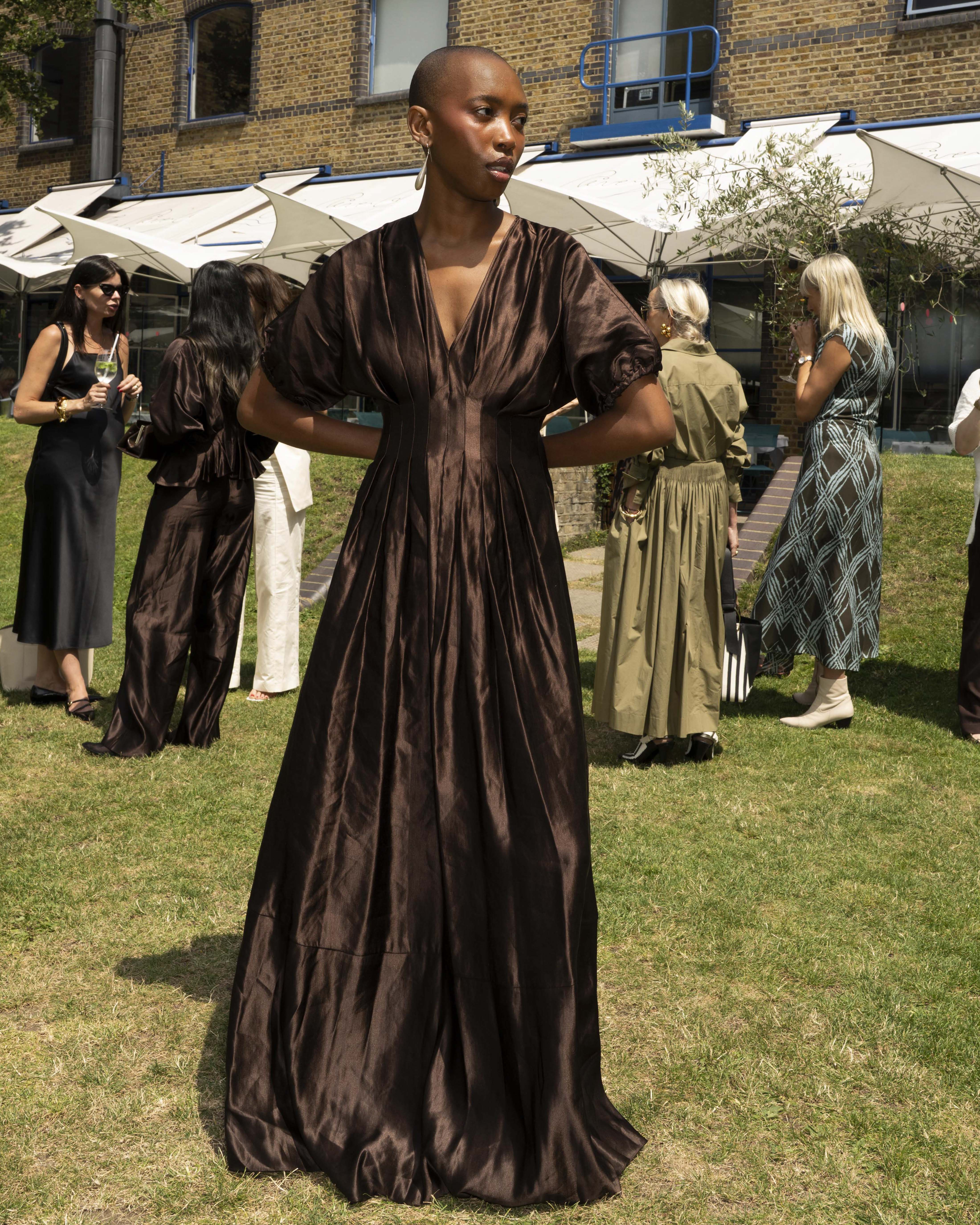 Marisa Martins wearing a dark brown satin maxi dress with puff sleeves and a deep V-neckline, at a summer garden event.