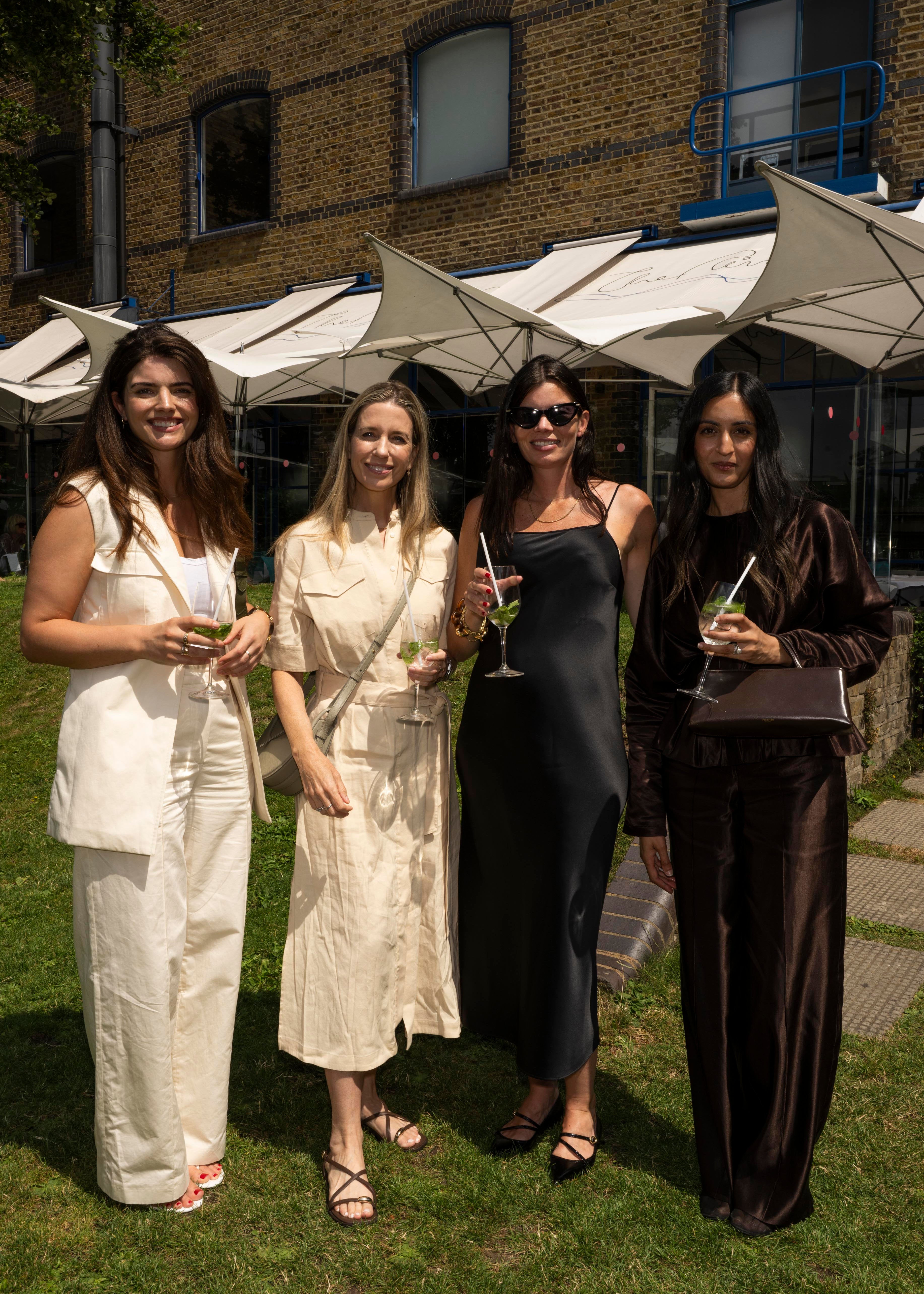 Ellie Stacey, Sarah Chapman, Ele Prideaux, Ume Romaan posing together at a summer event. They are dressed in stylish, coordinated outfits in neutral and dark tones, holding cocktails.