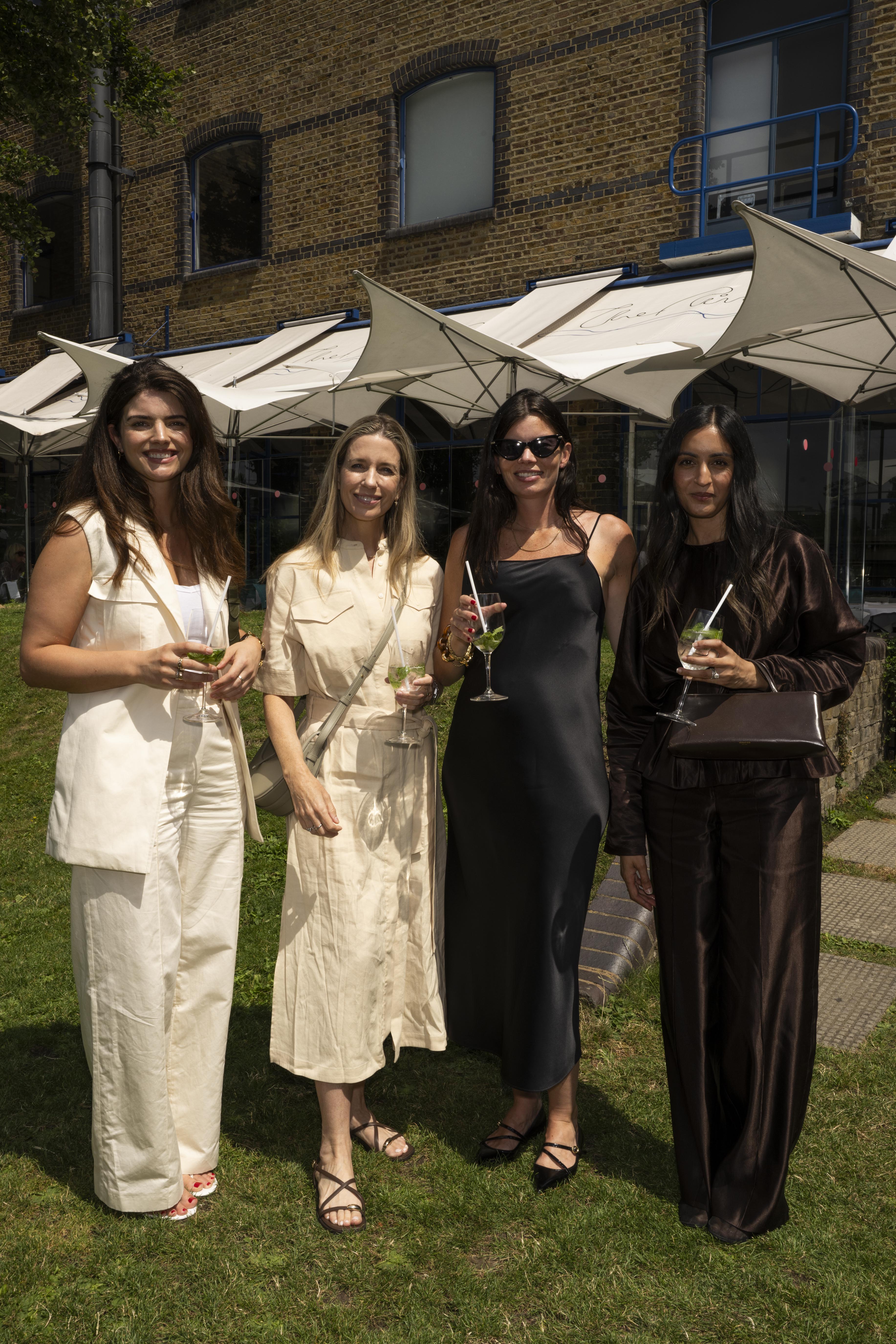 Ellie Stacey, Sarah Chapman, Ele Prideaux, Ume Romaan posing together at a summer event. They are dressed in stylish, coordinated outfits in neutral and dark tones, holding cocktails.