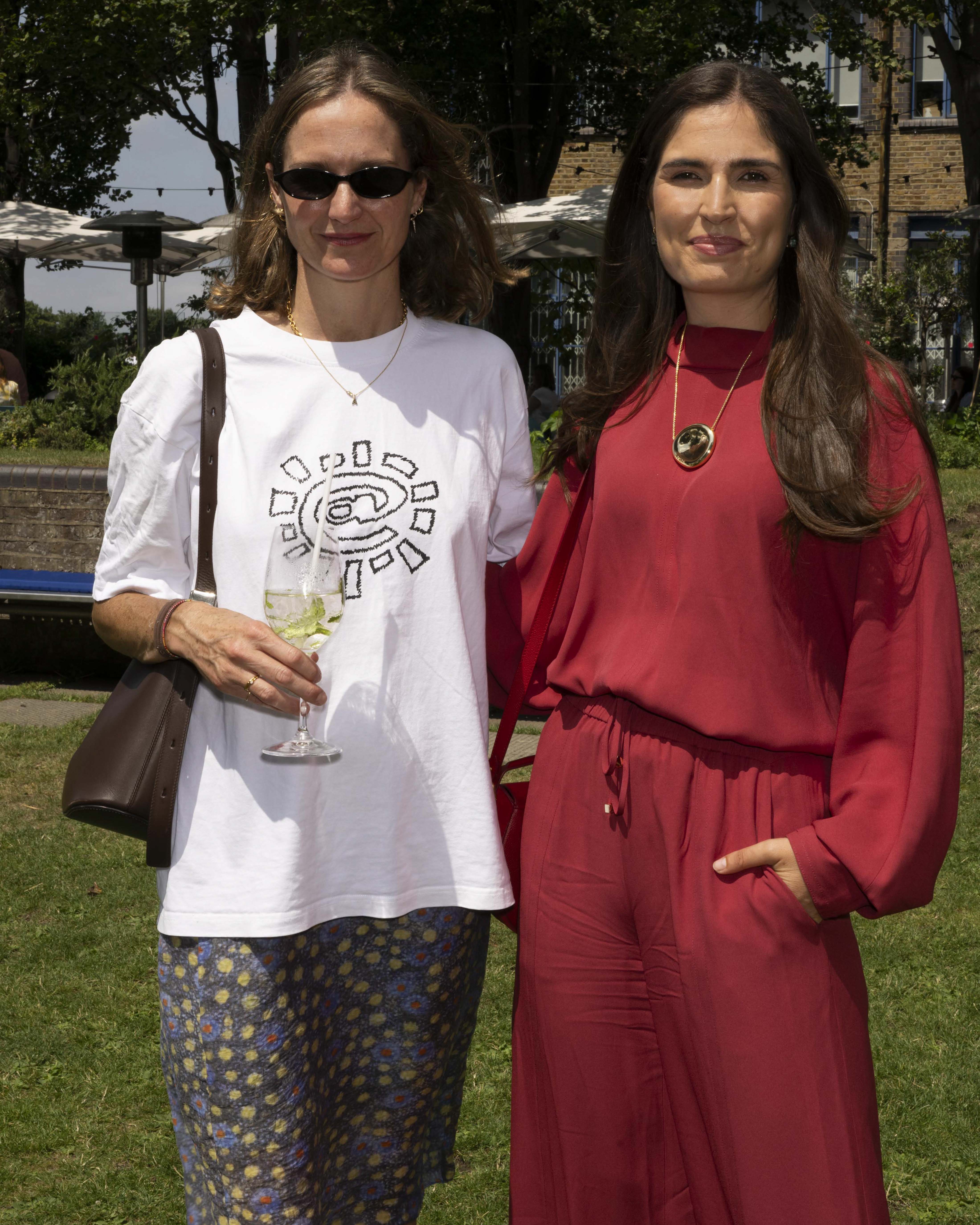 Elizabeth Sims, Marine Tanguy standing outdoors at a summer event. The woman on the left wears a white graphic T-shirt, patterned skirt, sunglasses, and carries a brown shoulder bag while holding a drink.
